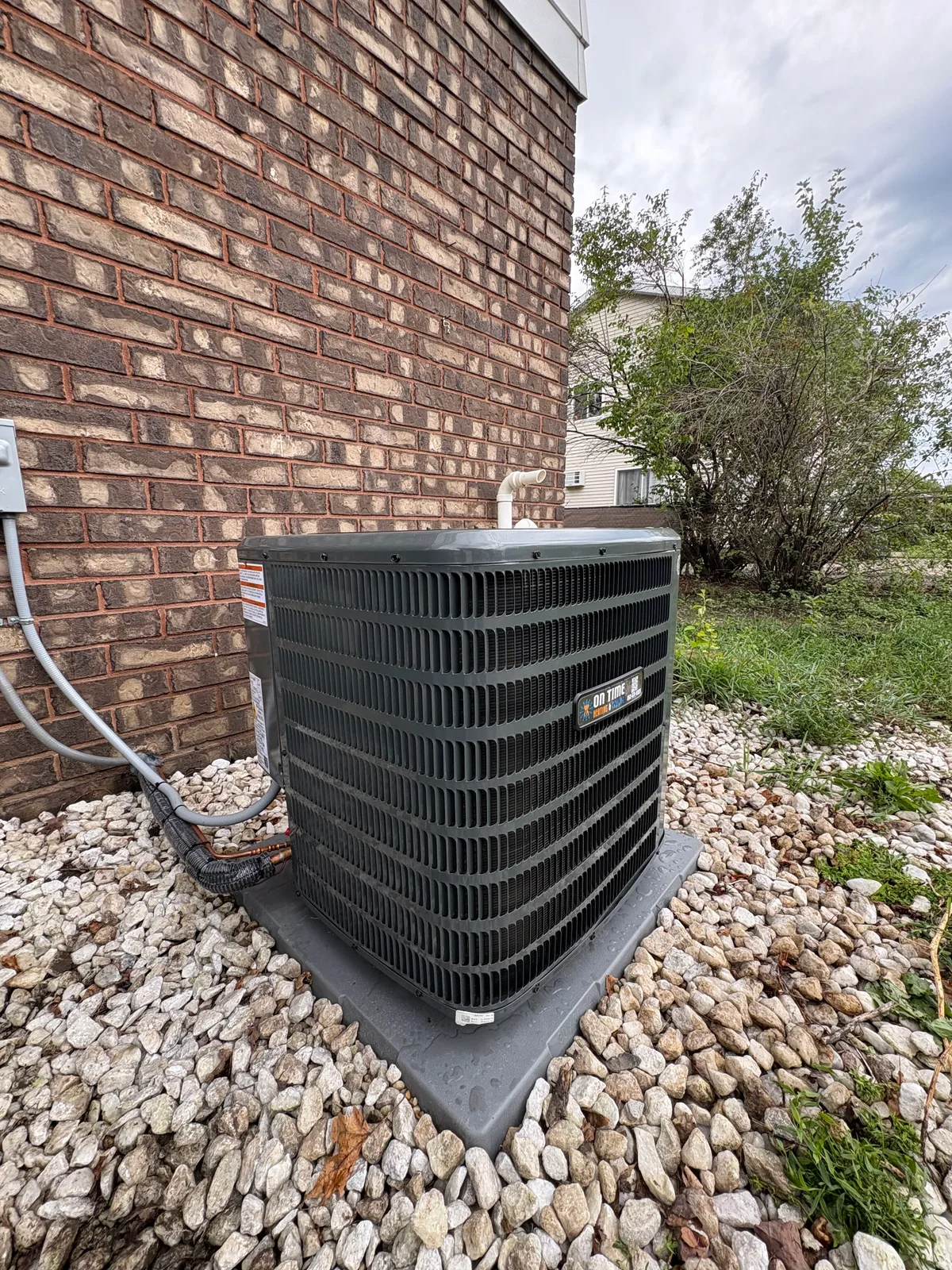 On Time Heating & Cooling air conditioner installed on a pad against a brick wall with gravel