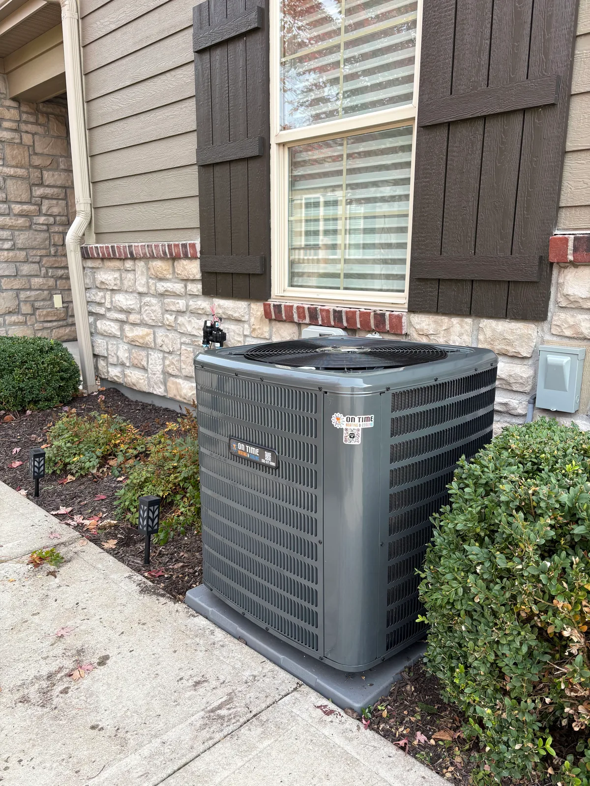 On Time Heating & Cooling air conditioner installed on a pad beside a stone home with shutters